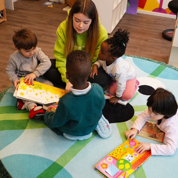 Eine Sprachförderkraft sitzt mit einer Gruppe Kinder auf dem Boden und liest ein Buch in der Kita kinderzimmer Hamburg