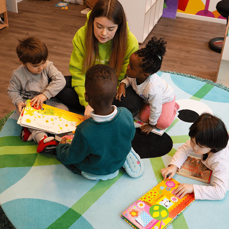Eine Sprachförderkraft sitzt mit einer Gruppe Kinder auf dem Boden und liest ein Buch in der Kita kinderzimmer Hamburg